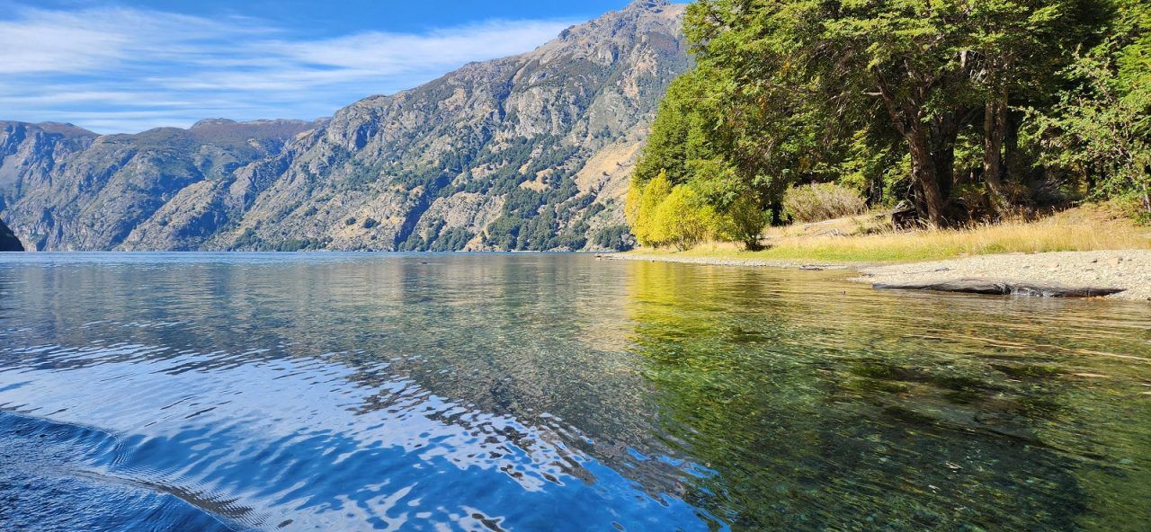 Clear, calm waters of Lago Azul lapping at the shore of the Coyhaique lot, with mountain views.